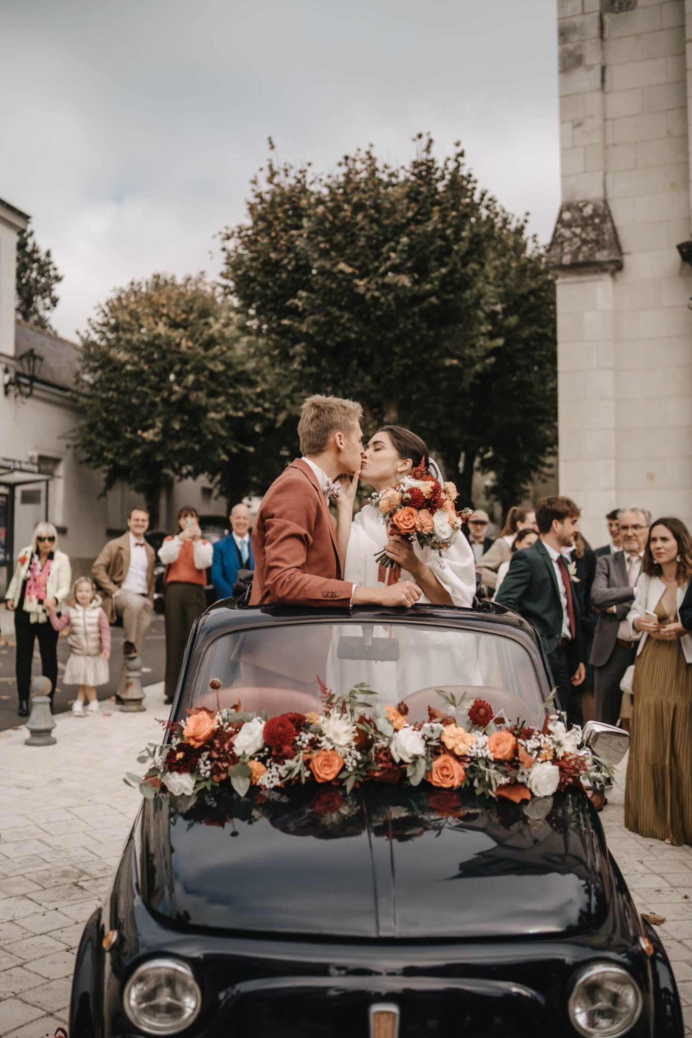 mariés qui s’embrassent dans une voiture ancienne décorée de fleurs à la sortie de la cérémonie